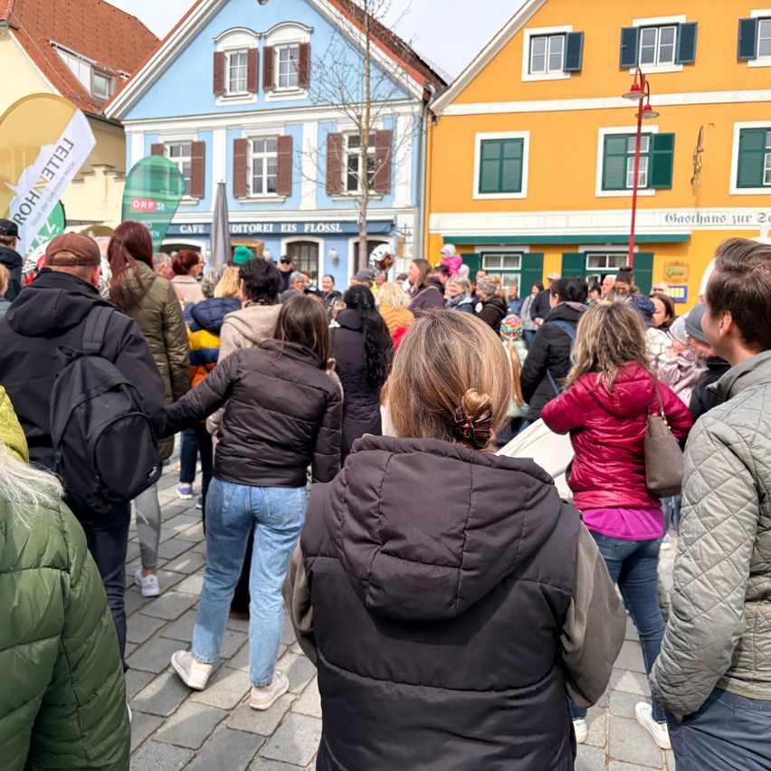 Eine Menschenmenge versammelt sich vor bunten Gebäuden, einige in Winterkleidung und Mützen, mit Bannern und einer Straßenlaterne sichtbar.
