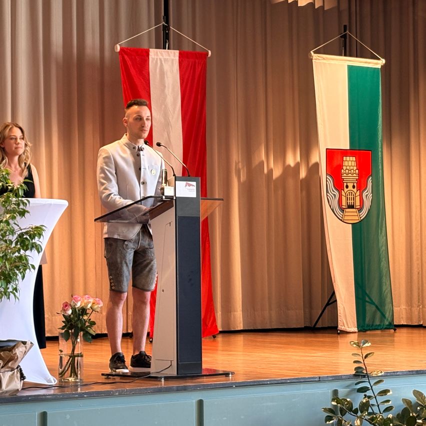 A man stands at a podium on stage, addressing the audience. Behind him, two flags are visible, one with a red and white design and the other green and white. A woman stands off to the side with a plant.