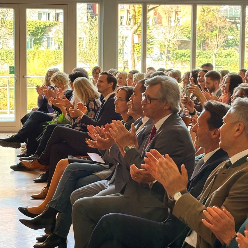 A crowd of formally dressed individuals are clapping in a room with large windows and a view of trees and buildings outside.