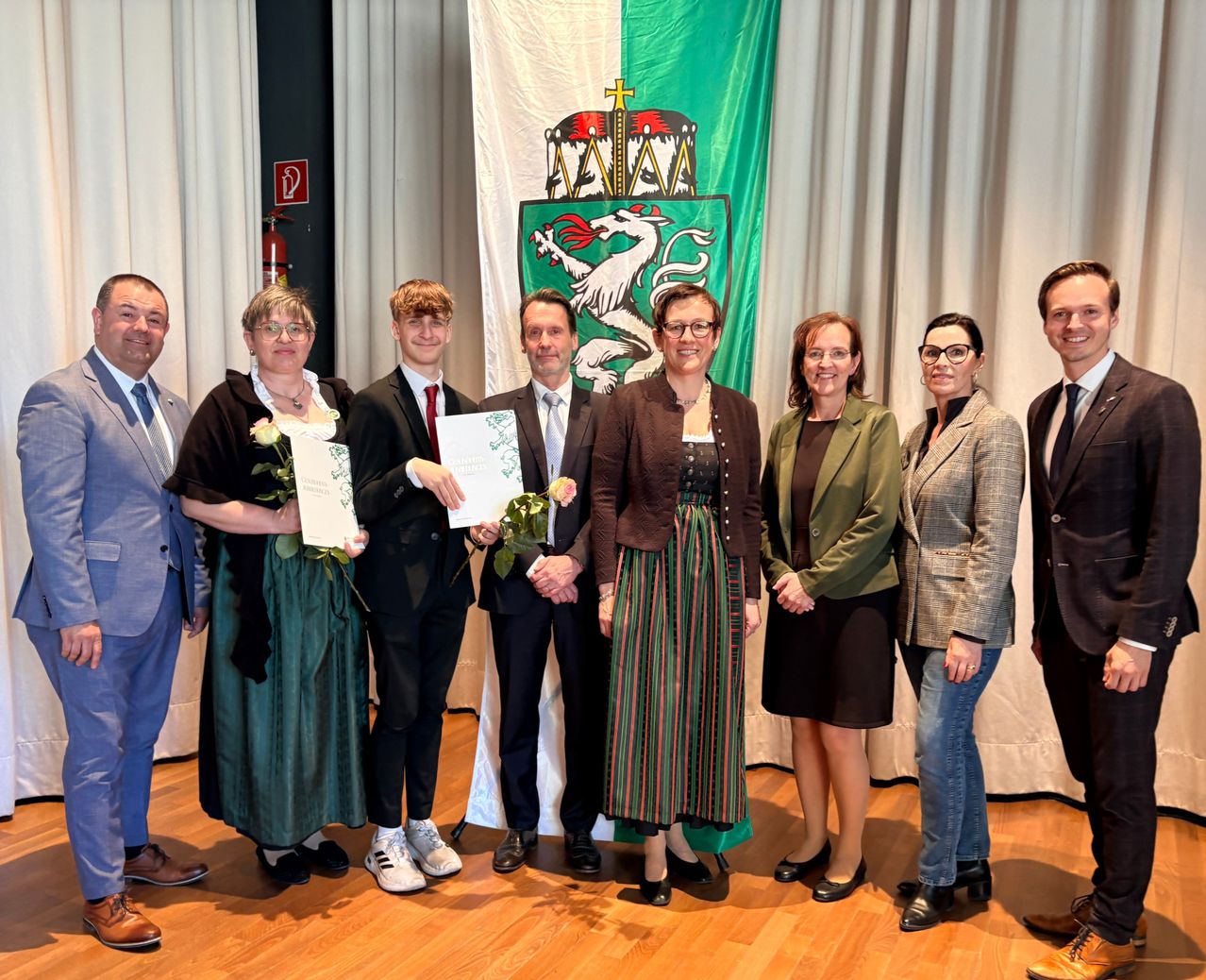 A group of individuals are standing in front of a flag with a white backdrop, holding certificates and flowers. They are all dressed formally.