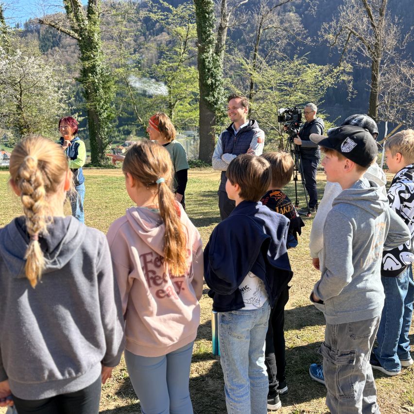 A group of children and adults gather in a park, surrounded by trees and mountains. One man is filming with a camera.
