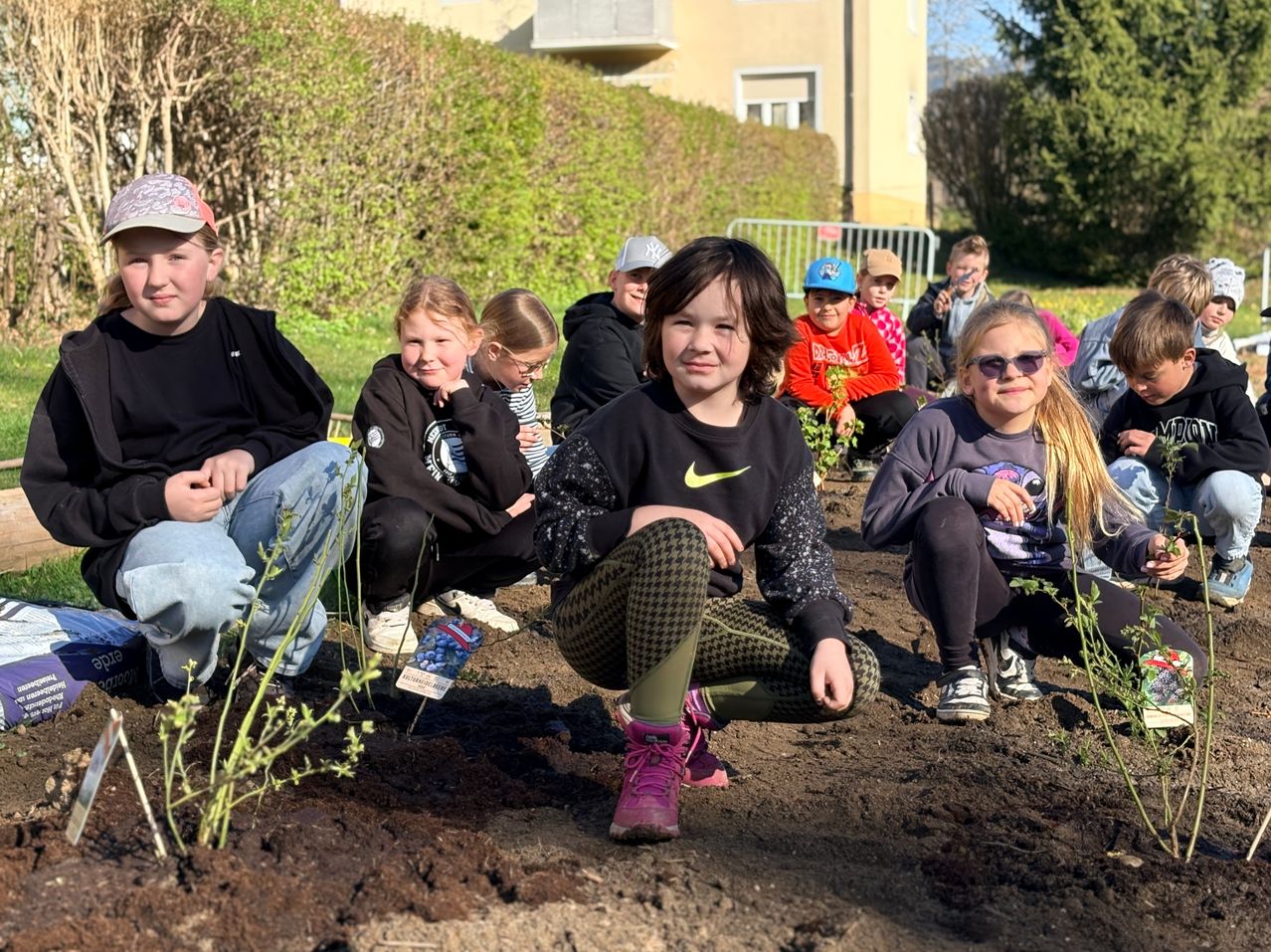 A group of children sit on the ground in a garden, wearing various clothing. They are surrounded by plants and soil, and a few adults are present.