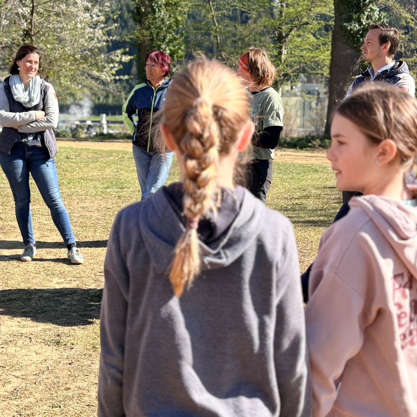 A group of adults and children stand outdoors, perhaps in a park, with one woman smiling and another with her arms crossed.