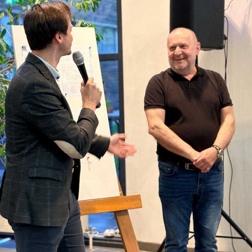 A man in a suit speaks into a microphone near a whiteboard, while another man in a brown shirt smiles. Behind them is a speaker on a stand.