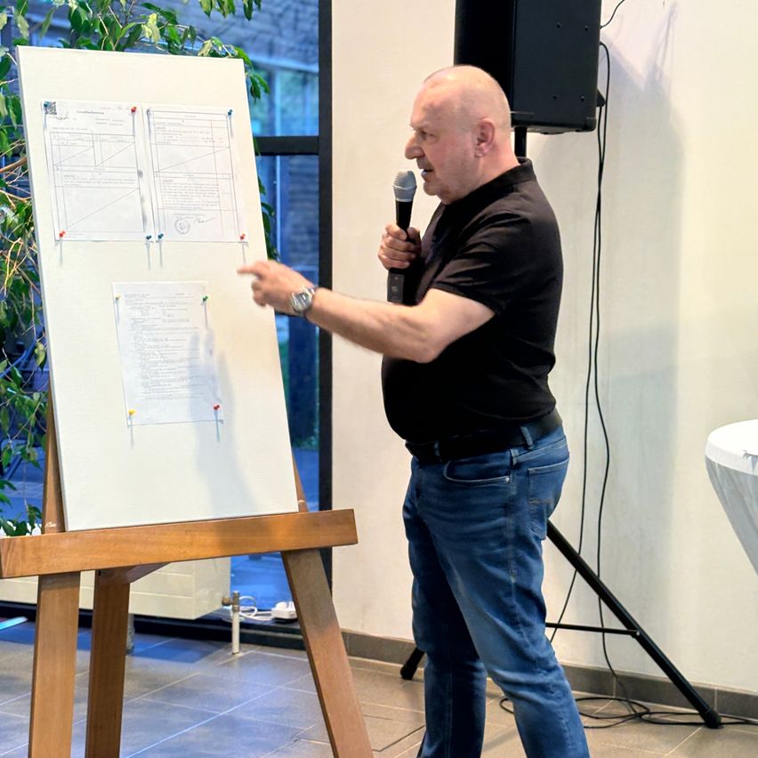 A man with a microphone stands in front of a flip chart and points at it with his right hand.