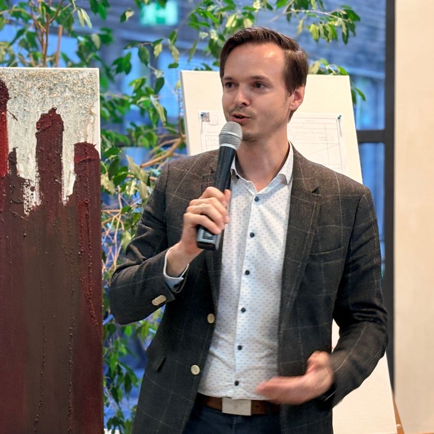 A man in a suit speaks into a microphone in front of a painting and a whiteboard.