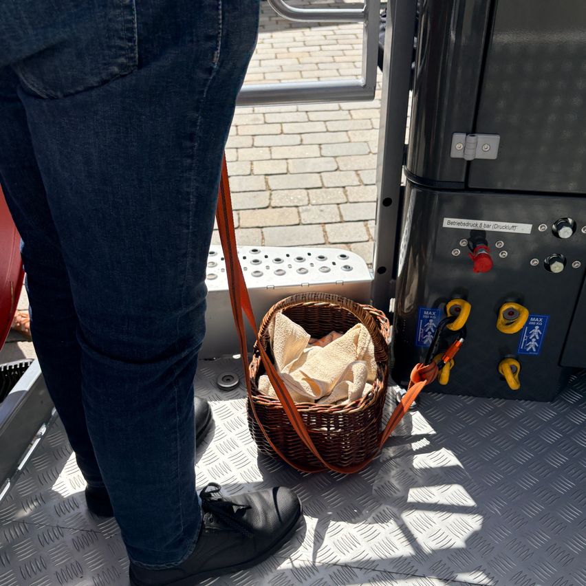 Eine Person in Jeans und schwarzen Schuhen hält einen Korb voller Handtücher auf einem Metallboden. In der Nähe ist ein schwarzes Objekt mit einem roten Knopf und mehreren gelben Griffen zu sehen.