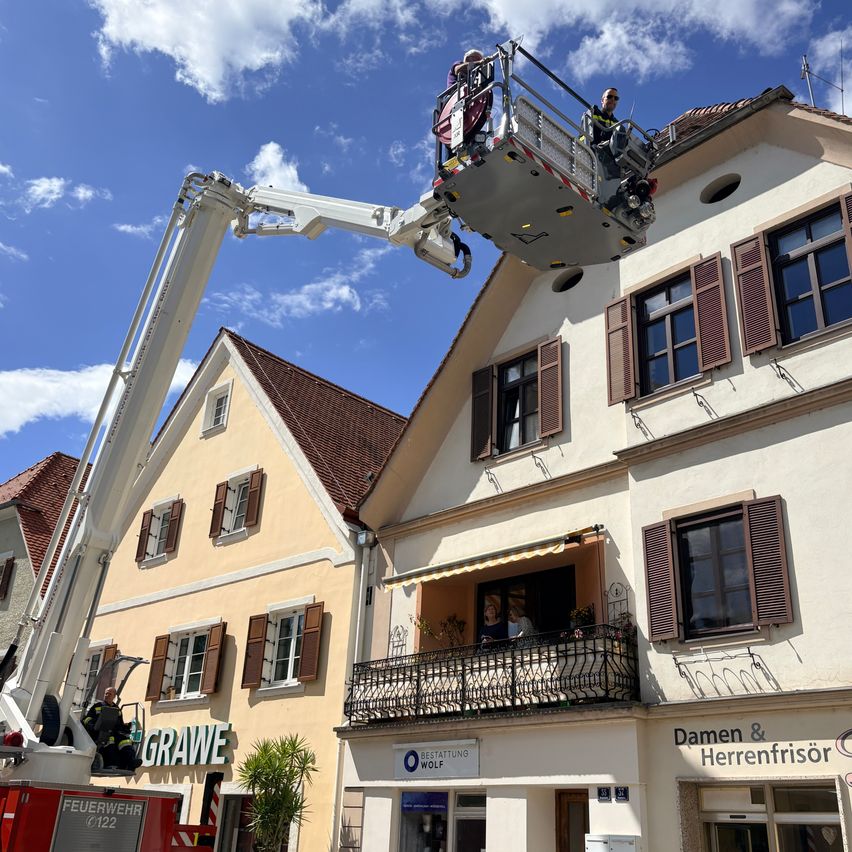 Feuerwehrleute benutzen eine Kran, um jemandem auf dem Dach eines Gebäudes zu helfen. Unten hat ein Balkon Personen, die herausschauen. Das Gebäude hat Schilder mit 'Damien & Herrenfrisor' und 'Feuerwehr'.