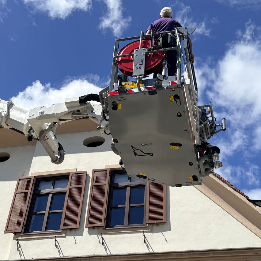Ein Mann steht auf einer Hebebühne und arbeitet an einem Gebäude mit drei Fenstern und einem roten Werkzeug. Der Himmel ist blau mit Wolken.