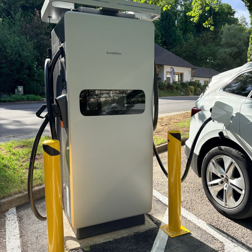 Ein weißer Elektrowagen lädt an einer Sungrow-Ladestation. Die Station hat ein Solarpanel oben und ist von gelben Pfosten umgeben. Bäume und ein Haus sind im Hintergrund.