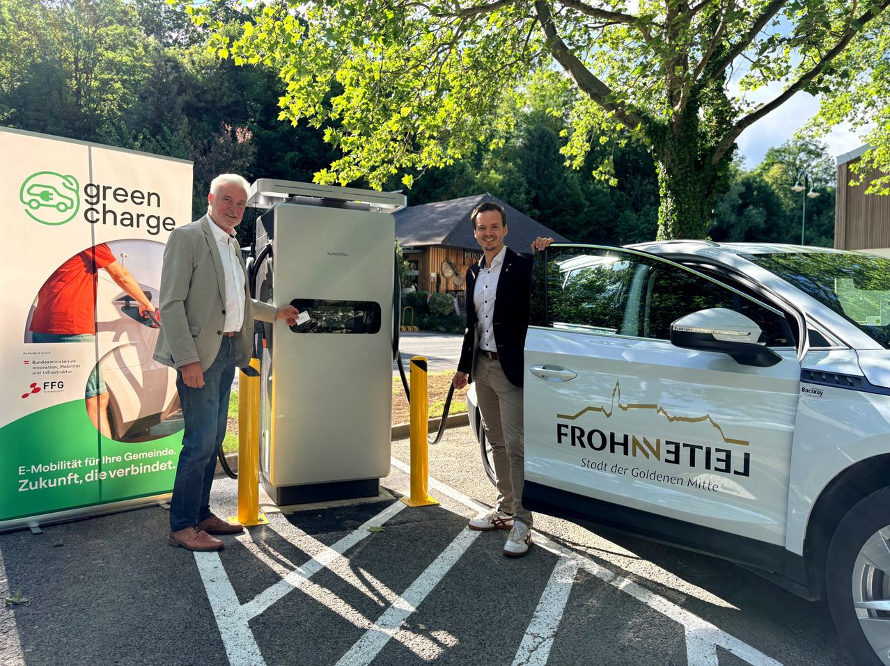 Zwei Männer stehen neben einer Ladestation für Elektroautos und lächeln. Einer hält ein Ladekabel. Ein Banner hinter ihnen sagt 'een charge'. Das Auto hat 'FROHNLEIEN' auf seiner Tür.