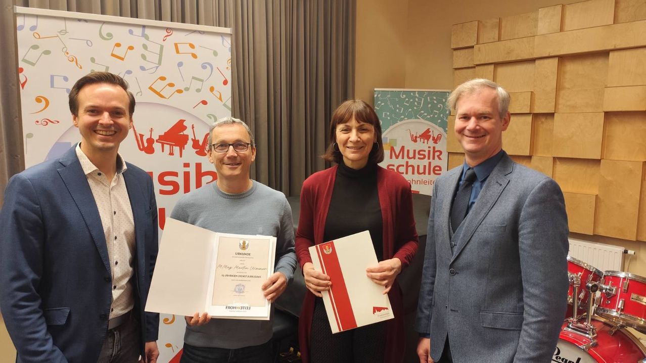 Three people are standing next to each other, holding certificates. The man on the left has a certificate for May Martin Winner. The woman in the middle has a certificate with a red and white cover. The man on the right is smiling and holding a certificate. Behind them, there are banners with musical notes and the word 'Musikschule'.