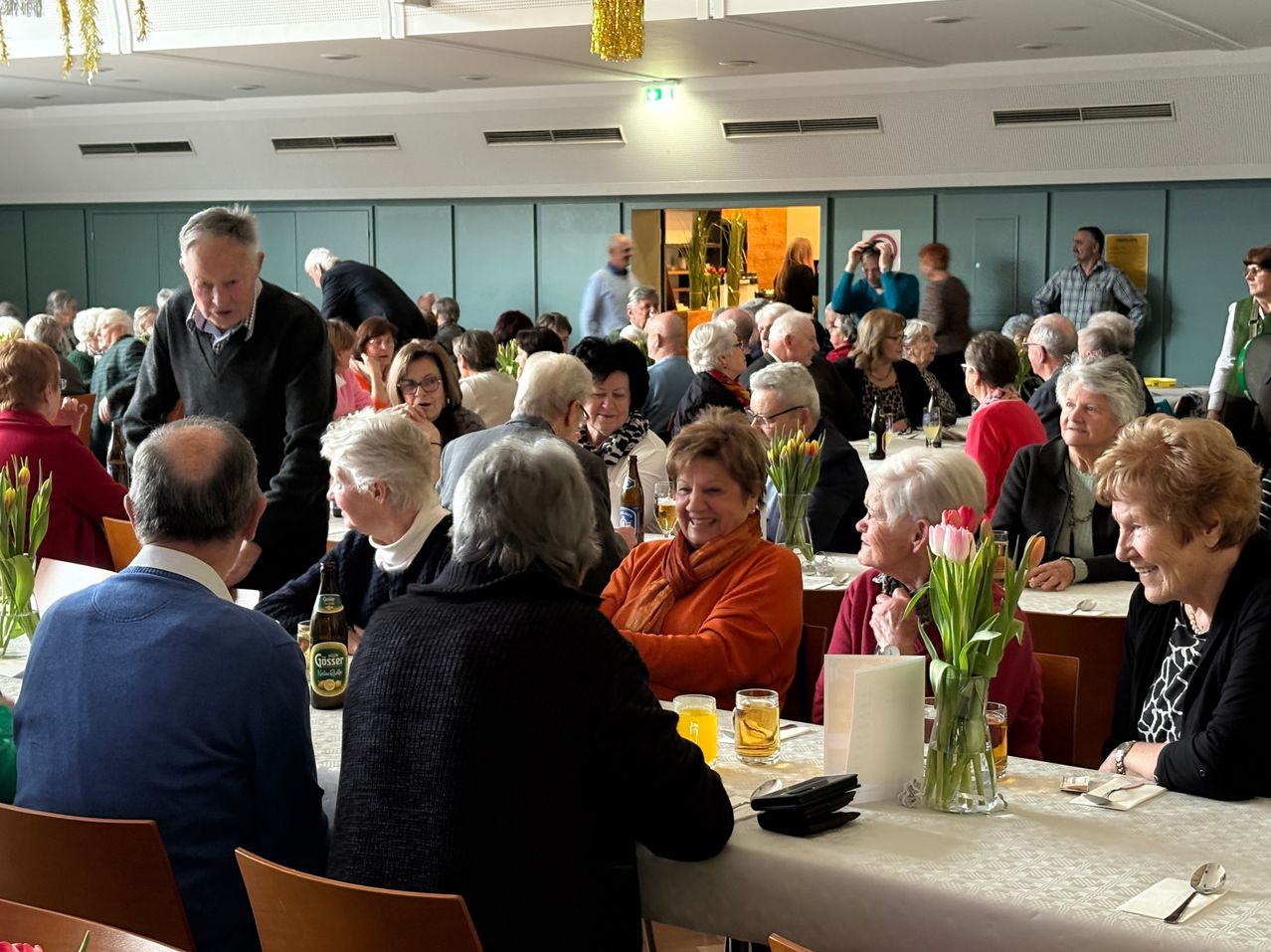 A group of elderly people sit at tables in a restaurant, some with glasses of beer and wine. A woman with a scarf smiles and a man in a black sweater looks at her.