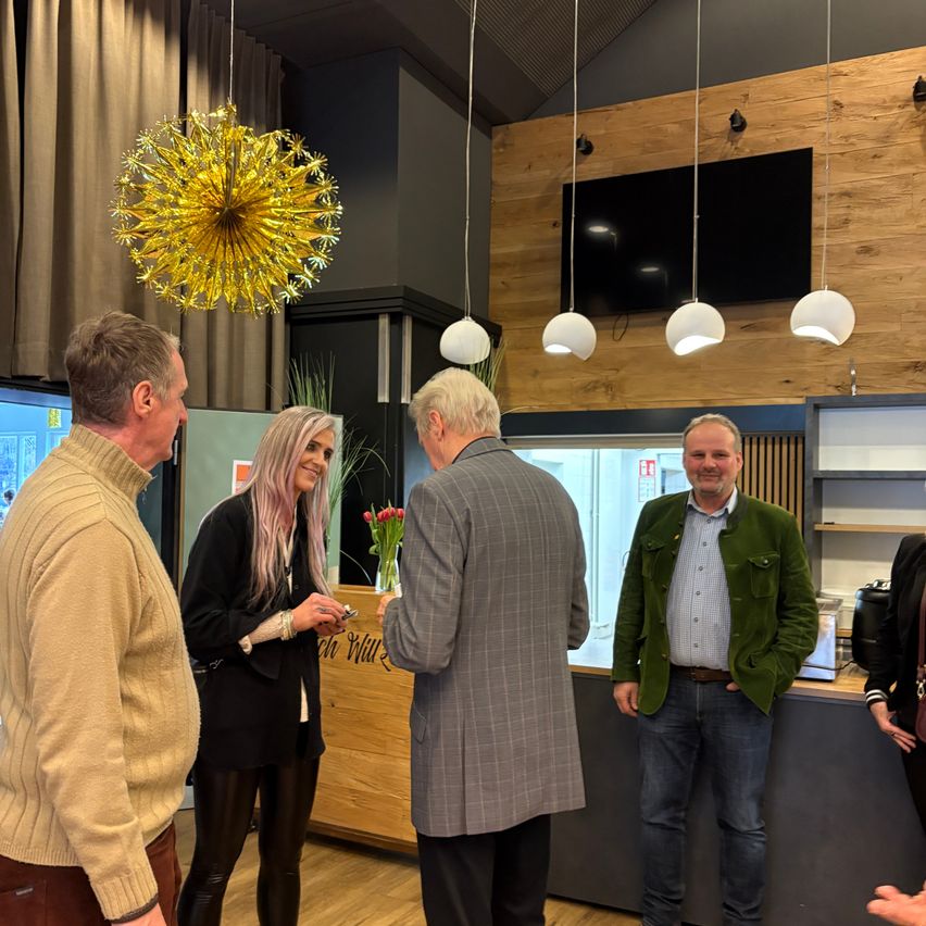 A group of people stand in a modern room. A woman hands a card to a man. A decorative chandelier and a TV are in the background.