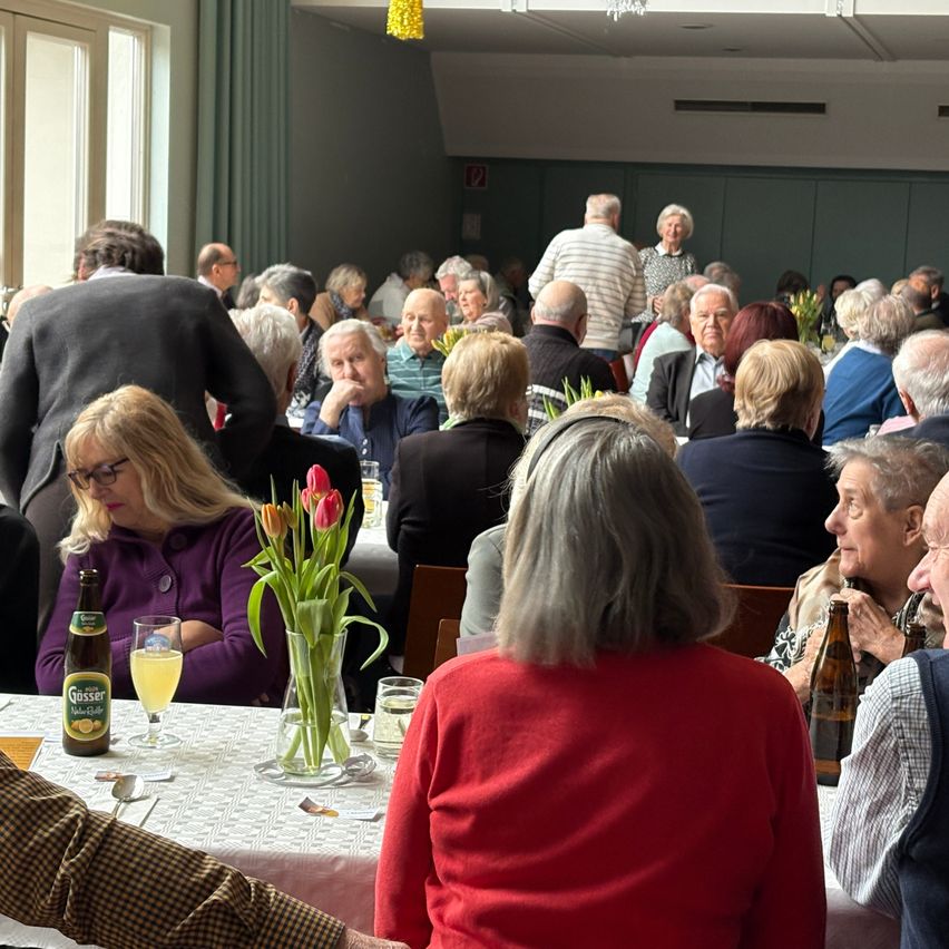 A group of people sit around tables in a dining hall, engaged in conversation. A woman in purple sits next to a vase of tulips, while others have drinks.