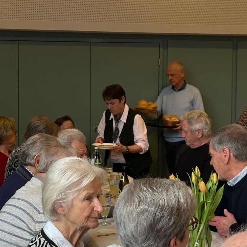 A group of older adults sit around a table, engaged in conversation. One woman holds a plate of bread, while another holds a glass of wine. A man with glasses stands nearby.