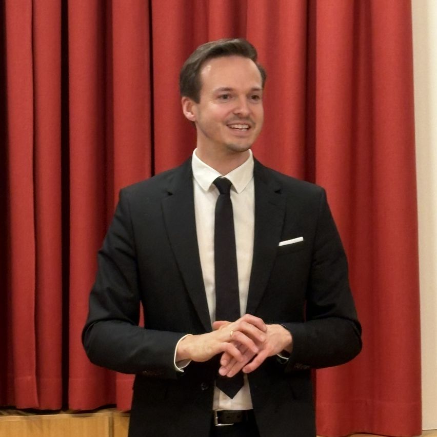 A man stands in front of a red curtain, dressed in a black suit, white shirt, and black tie. He smiles and clasps his hands together.