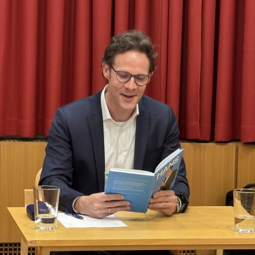 A man in a suit reads a book titled 'Hans' at a table, with glasses, a pen, and two glasses of water in front of him. Behind him is a red curtain.