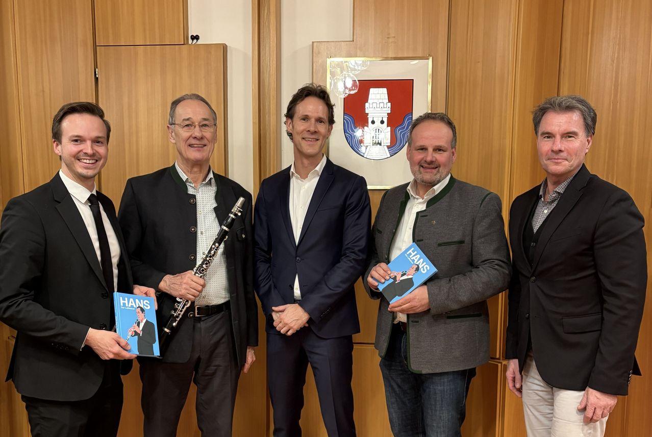 Five men stand together in a room, all holding books. The man on the left holds a clarinet. The middle man has a white shirt and dark suit. Behind them, a picture frame with a coat of arms is mounted on the wall.