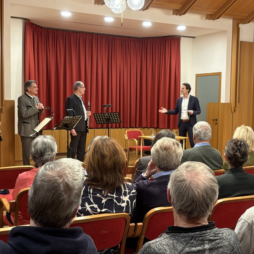 A music recital is taking place in a hall. Three musicians are standing on stage, two holding clarinets, and one gesturing. An audience of various ages is seated in front of them. The stage has a red curtain backdrop and a wooden floor.