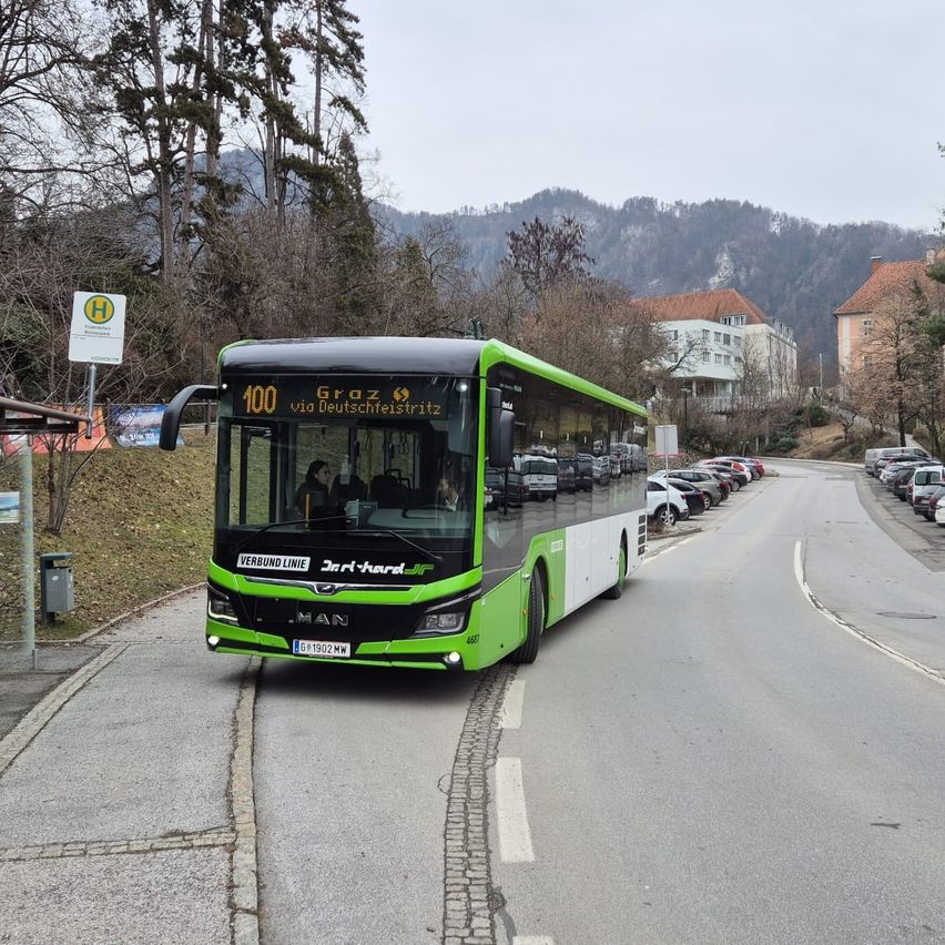 A green bus is driving on a road, with several people inside and parked cars on the side. A building and mountains are in the background.