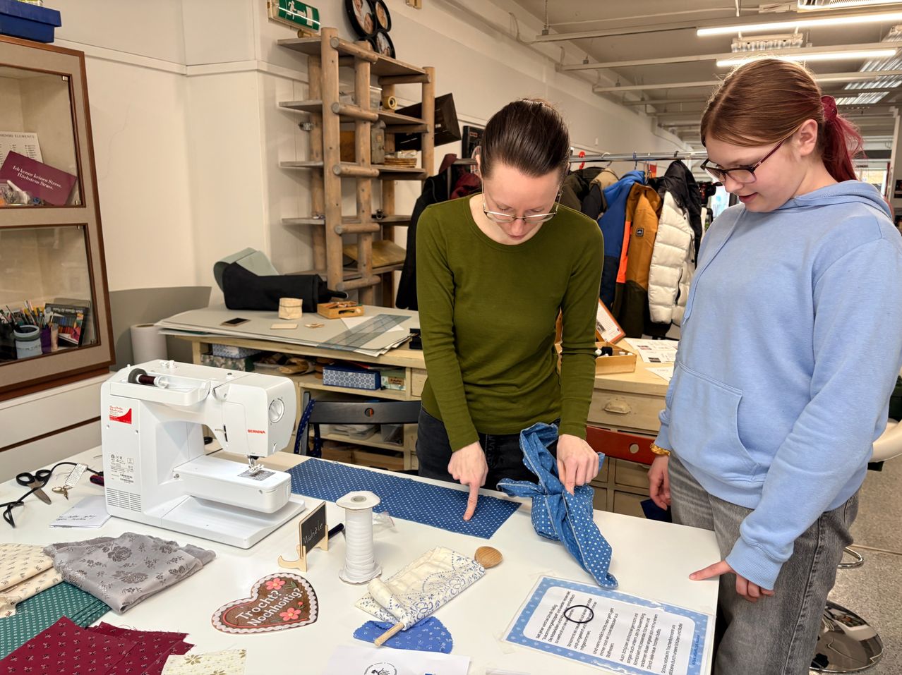 Two women are in a workshop, one is showing the other how to tie a tie. There is a sewing machine, spools of thread, and various materials on the table.
