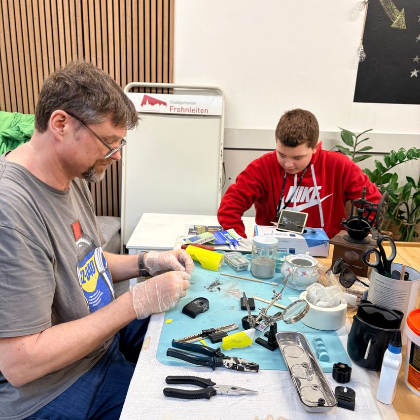 Two individuals work on a project at a table, surrounded by tools and materials. One man wears glasses and a gray shirt, while the other wears a red hoodie.