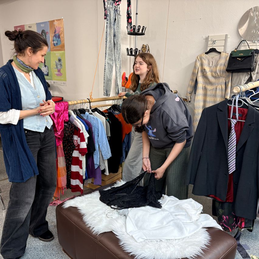 Three women in a clothing store, one is inspecting clothes, another is folding them, and the third is looking at a white garment on a fur cushion.