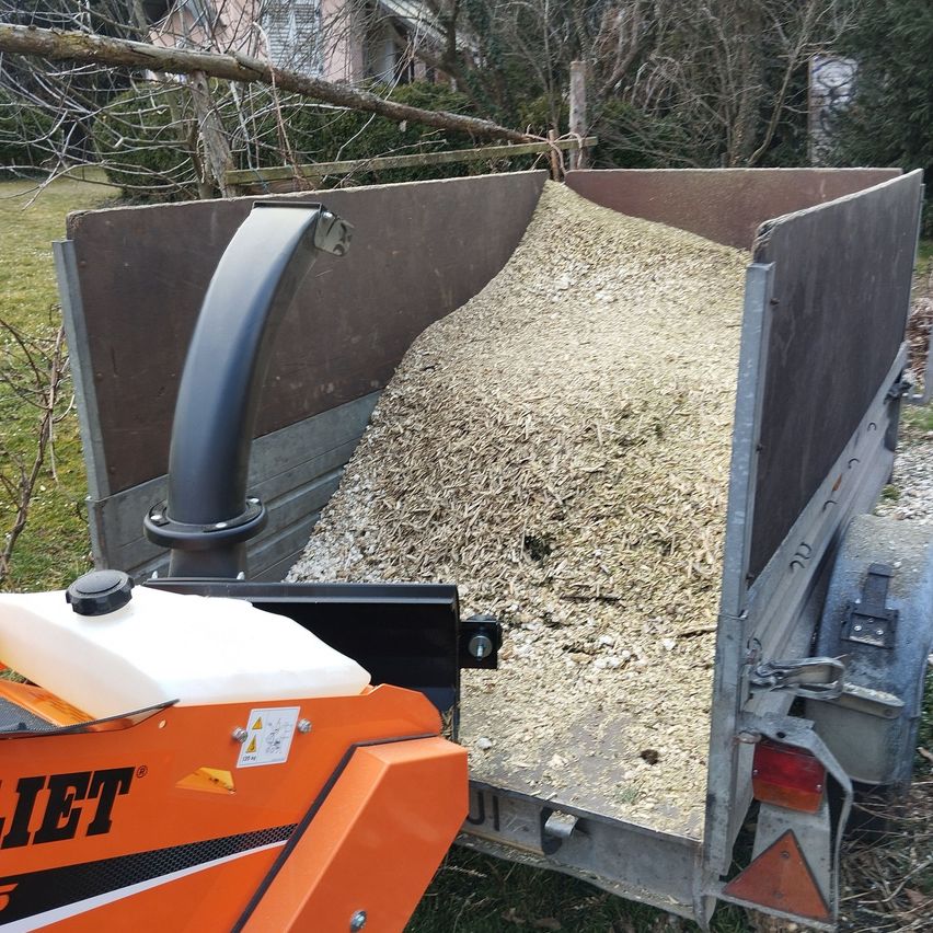 A truck is loaded with wood chips and is parked on grass. A machinery vehicle is attached to the truck.