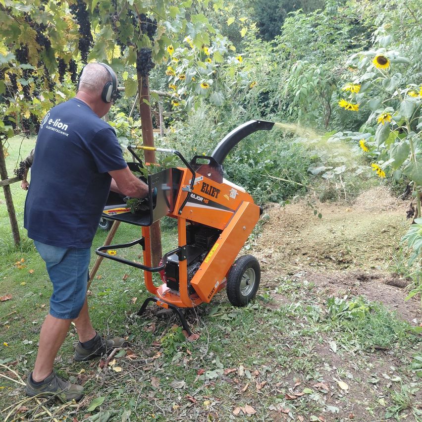 A man wearing headphones operates a large orange machine outdoors, surrounded by greenery.