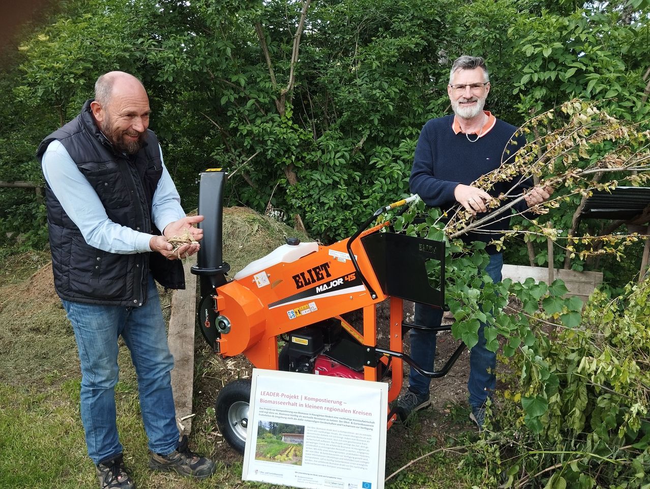 Two men are standing next to a machine outdoors. One man is holding some plant material and smiling. The other man is holding a branch and appears to be explaining something.