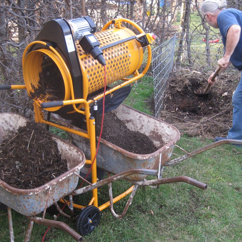 An older man digs the soil with a shovel near a yellow composter. There are two rusty wheelbarrows filled with compost beside him.