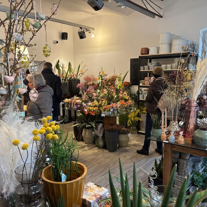 A flower shop filled with a variety of flowers in different colors and sizes. There are people browsing the flowers. The shop has wooden floors, white walls, and a shelf with decorative items.