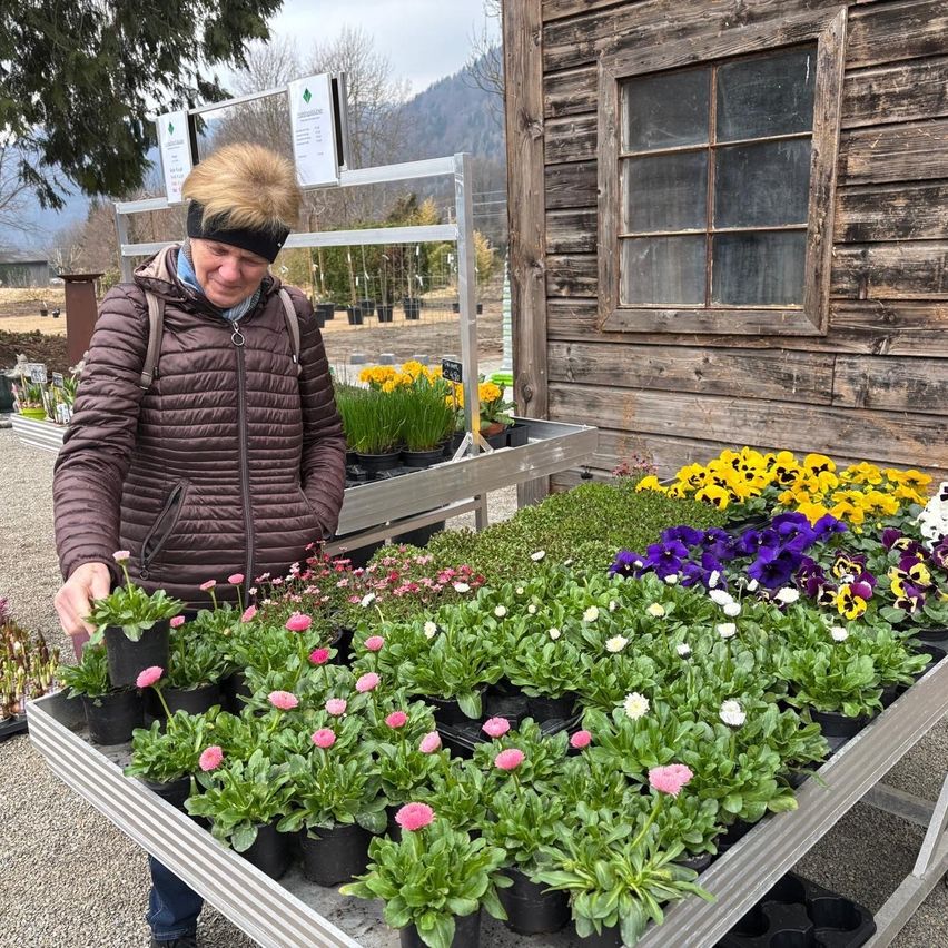 A woman stands outside a wooden building, smiling and looking at a large display of potted flowers.