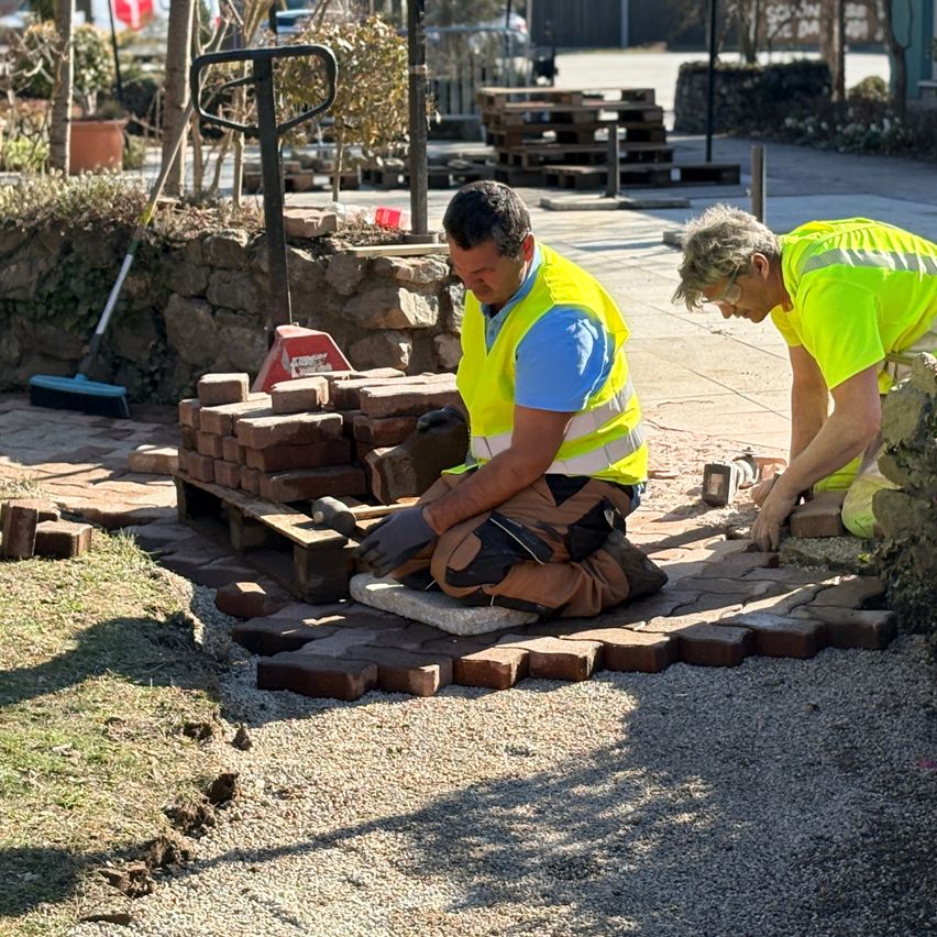 Two men in reflective vests are working on a brick pathway on a sunny day. One man is crouching while the other is standing. Nearby, there are stacks of bricks and a broom.