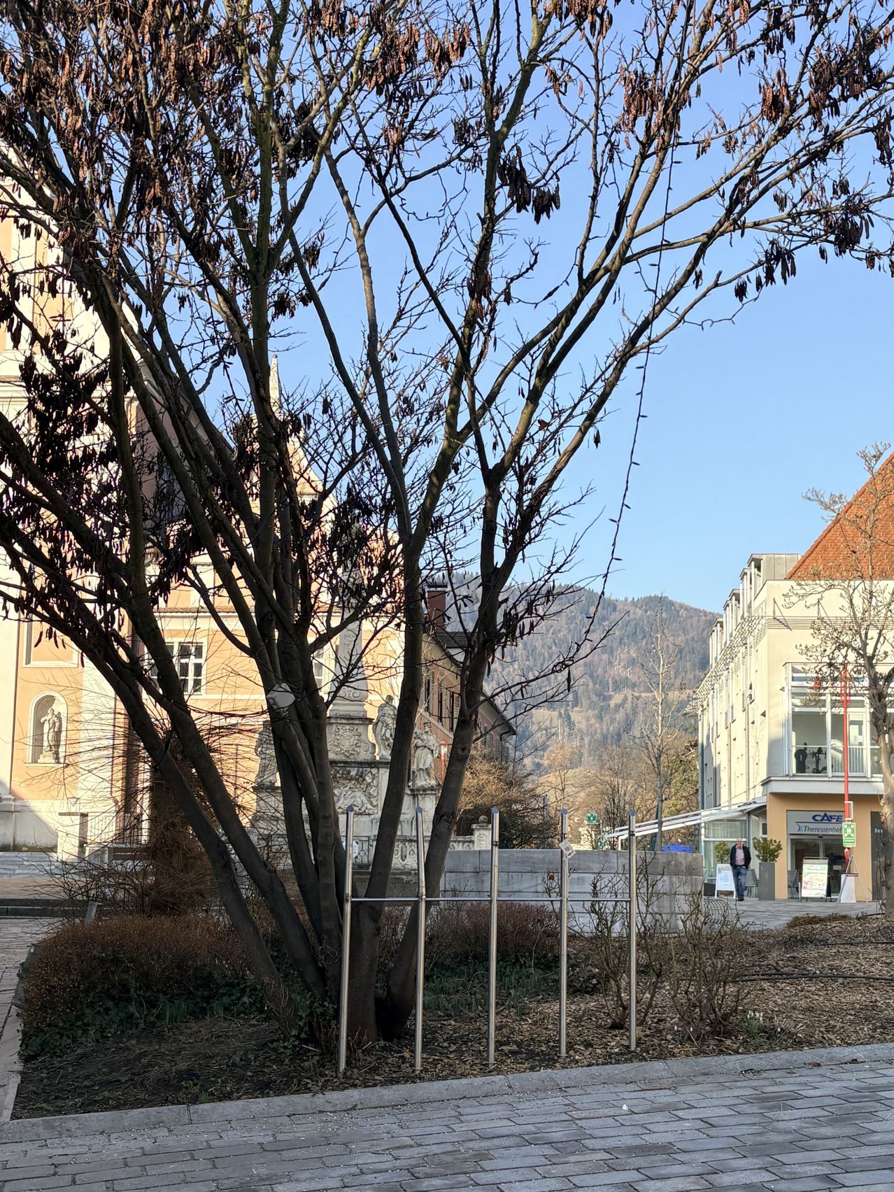 Ein Baum steht in einem Innenhof mit einem alten Gebäude dahinter, das Statuen und Fenster aufweist. Ein Café mit Balkon befindet sich rechts, ein Mensch geht vorbei. Berge sind im Hintergrund zu sehen.