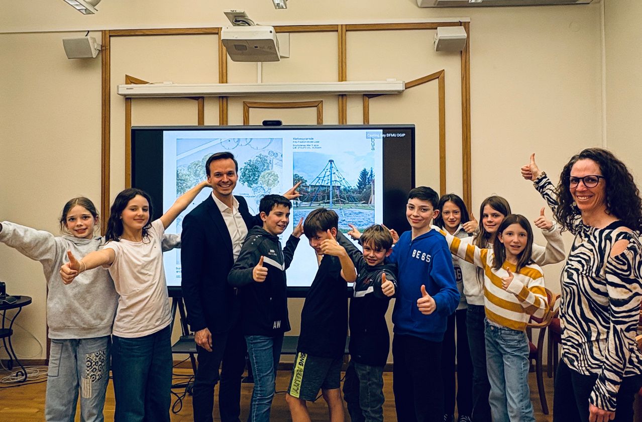 Eine Gruppe von Kindern und ein Mann stehen vor einer Projektionsleinwand, die einen Karussell zeigt. Sie alle zeigen Daumen hoch.