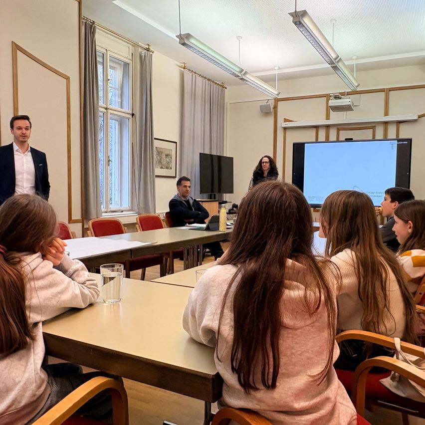 Ein Klassenzimmer mit Schülern, die aufmerksam einer Präsentation zuhören. Ein Mann steht vorne mit einer Projektionsleinwand dahinter. Ein anderer Mann sitzt an einem Schreibtisch, und mehrere Schüler sind an Tischen platziert.