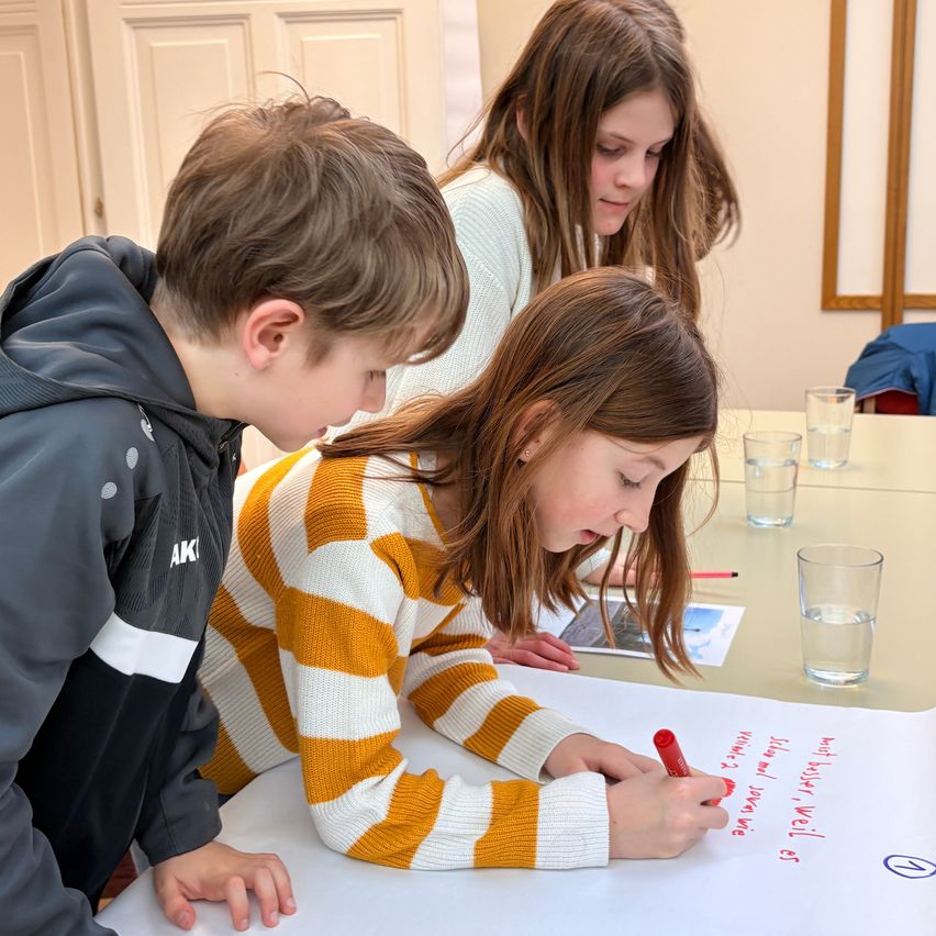 Drei Kinder sitzen an einem Tisch, ein Mädchen schreibt auf ein großes weißes Papier mit einem roten Marker. Ein anderes Mädchen schaut zu, und ein Junge blickt über das Papier. Dahinter stehen zwei Gläser Wasser und ein Foto auf dem Tisch.