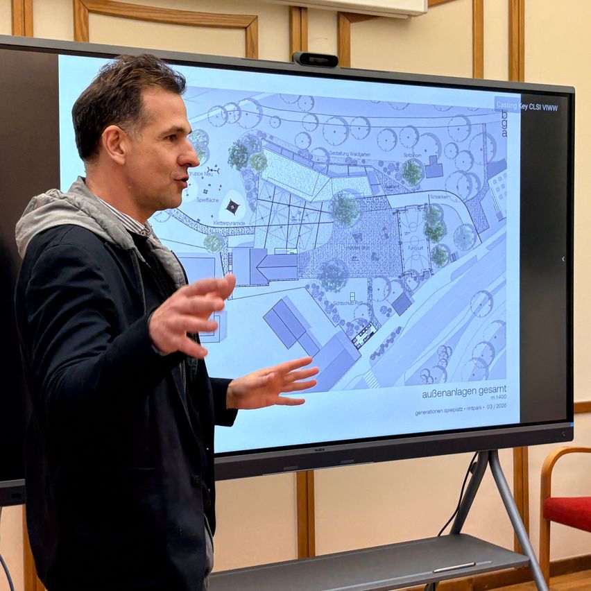 Ein Mann in einem schwarzen Mantel steht vor einer großen Leinwand und erklärt eine Landschaftsgestaltung. Die Leinwand zeigt eine detaillierte blaue Karte mit verschiedenen Pflanzen und Strukturen.