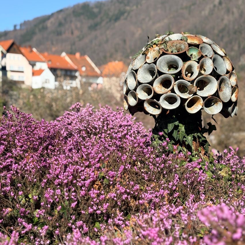 Ein Feld mit rosa Blumen und einer großen, pilzartigen Struktur aus Metall im Vordergrund. Im Hintergrund befinden sich Häuser mit orangefarbenen Dächern.