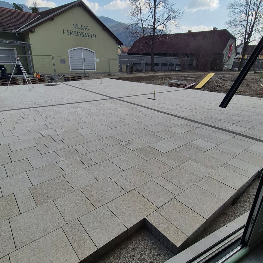 Ein Baustelle mit einem großen grauen Steinfliesenboden. Im Hintergrund ist ein Gebäude namens Musik-Vereinsheim zu sehen.