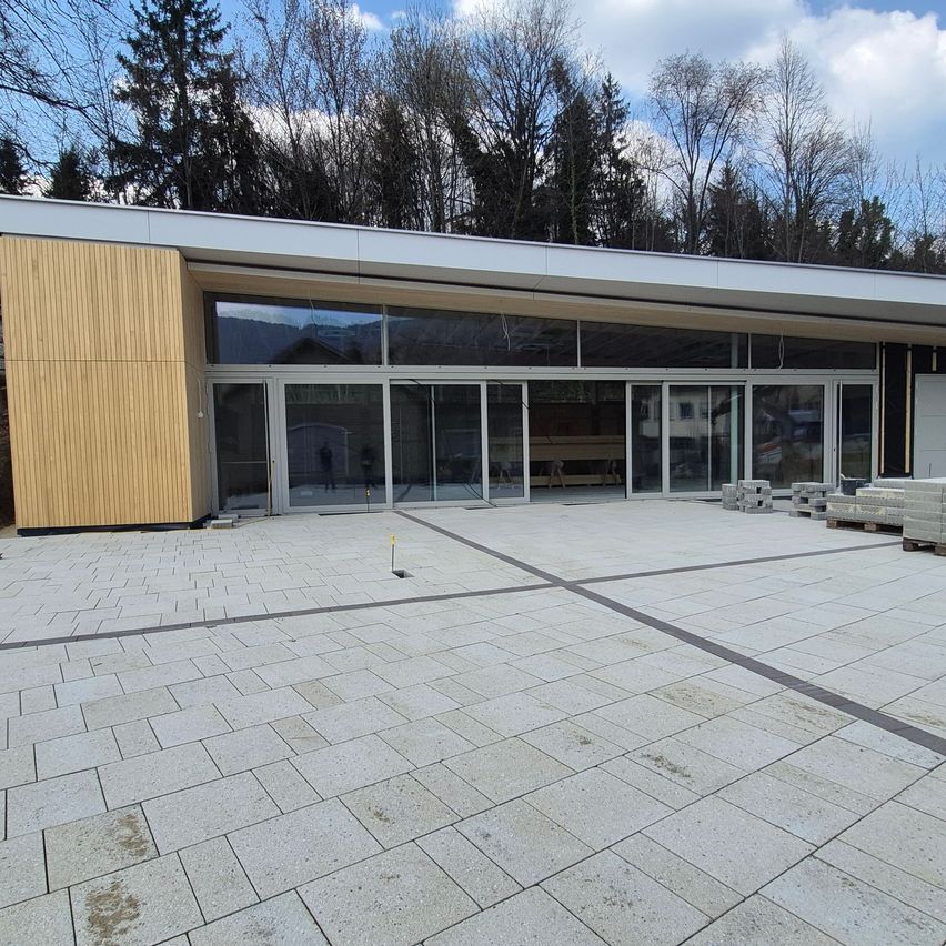 Modernes Gebäude mit Glaswänden und einer hölzernen Akzentwand, umgeben von Bäumen. Der Boden ist gefliest, und es gibt Baumaterialien in der Nähe.