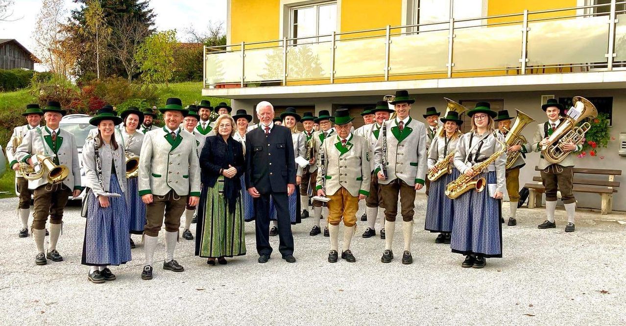 A group of musicians in traditional attire, including green hats and white coats, stand in front of a yellow building. A woman in a black coat and a man in a dark suit are in the center.