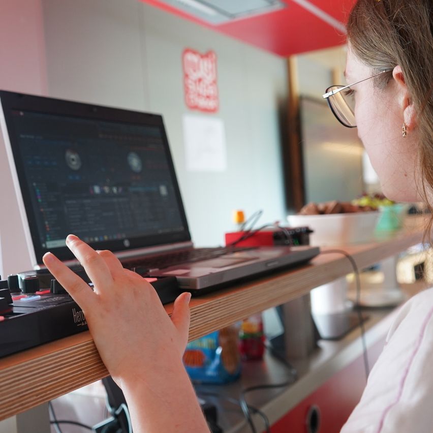Eine Frau mit Brille und weißem Shirt arbeitet an einem Laptop auf einem Holztisch. Daneben befindet sich ein schwarzer Controller.