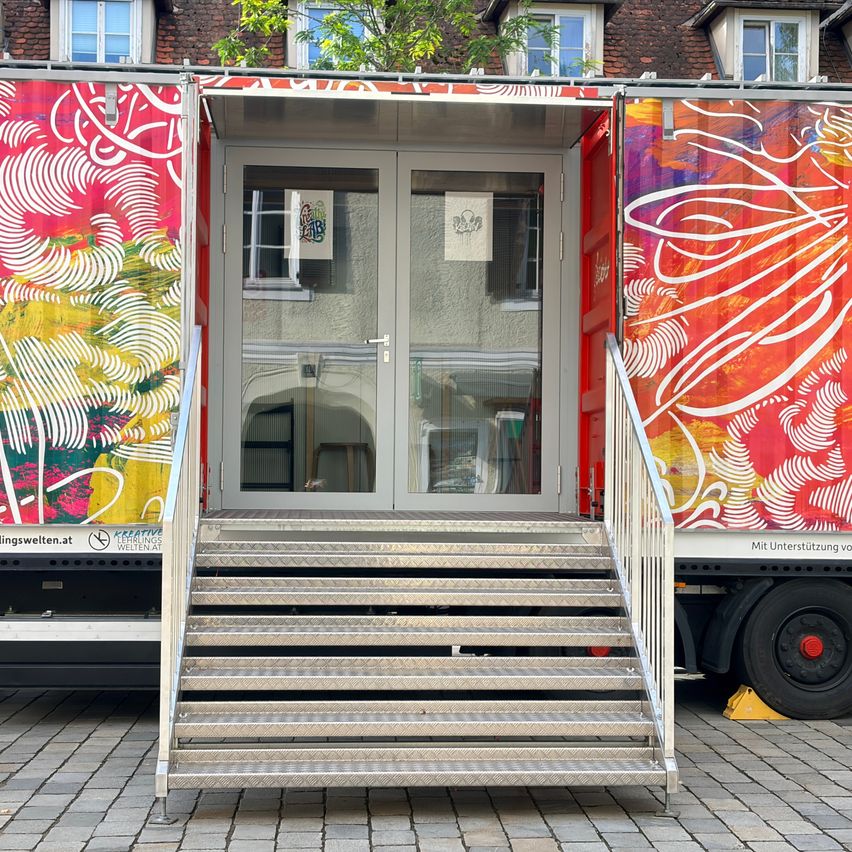 Ein farbenfroher Container mit einer Glastür und Metallstufen, die zu ihm hinaufführen, auf einem gepflasterten Bürgersteig geparkt.