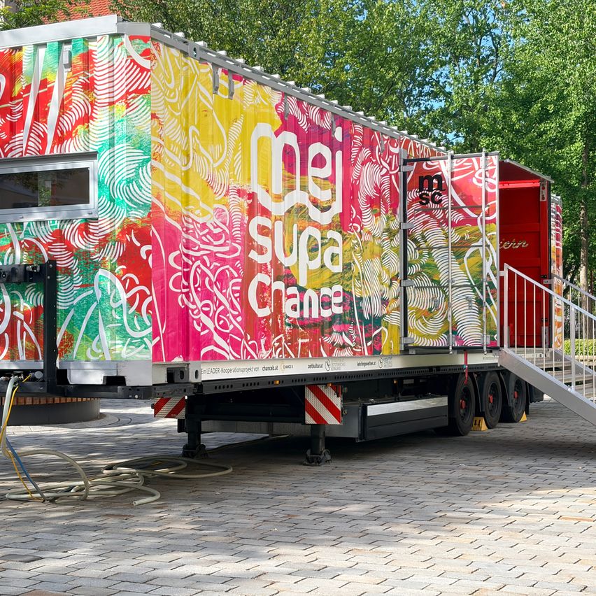 Ein farbenfroh bemalter Lastwagen mit 'mel supa chance' in weiß auf einem bunten Hintergrund, geparkt auf einer gepflasterten Oberfläche mit einer Metallrampe, die zur Tür führt.