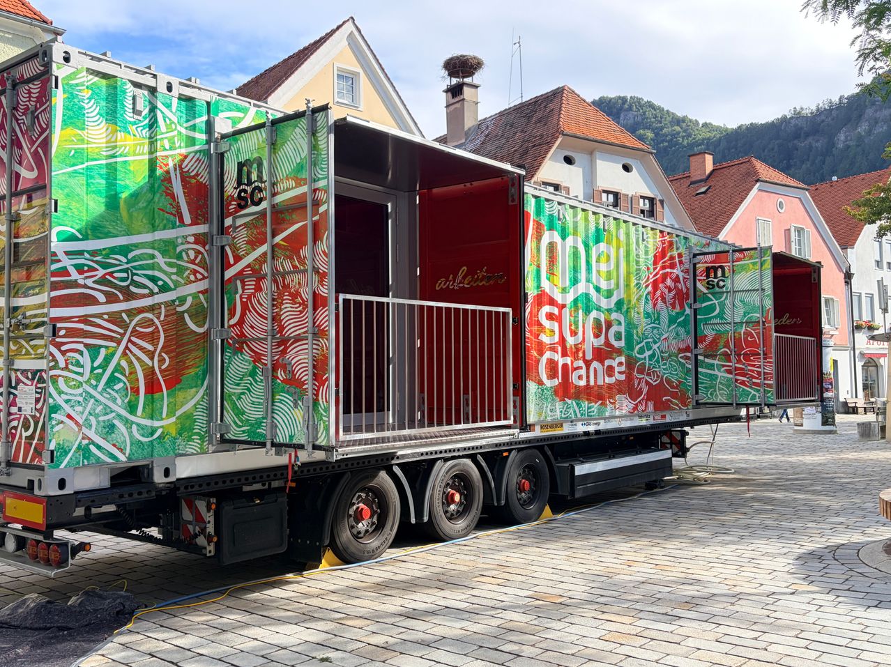Ein LKW mit einer roten Tür und bunten Graffitis steht auf einer Kopfsteinpflasterstraße vor Häusern mit Schornsteinen und Bergen im Hintergrund.