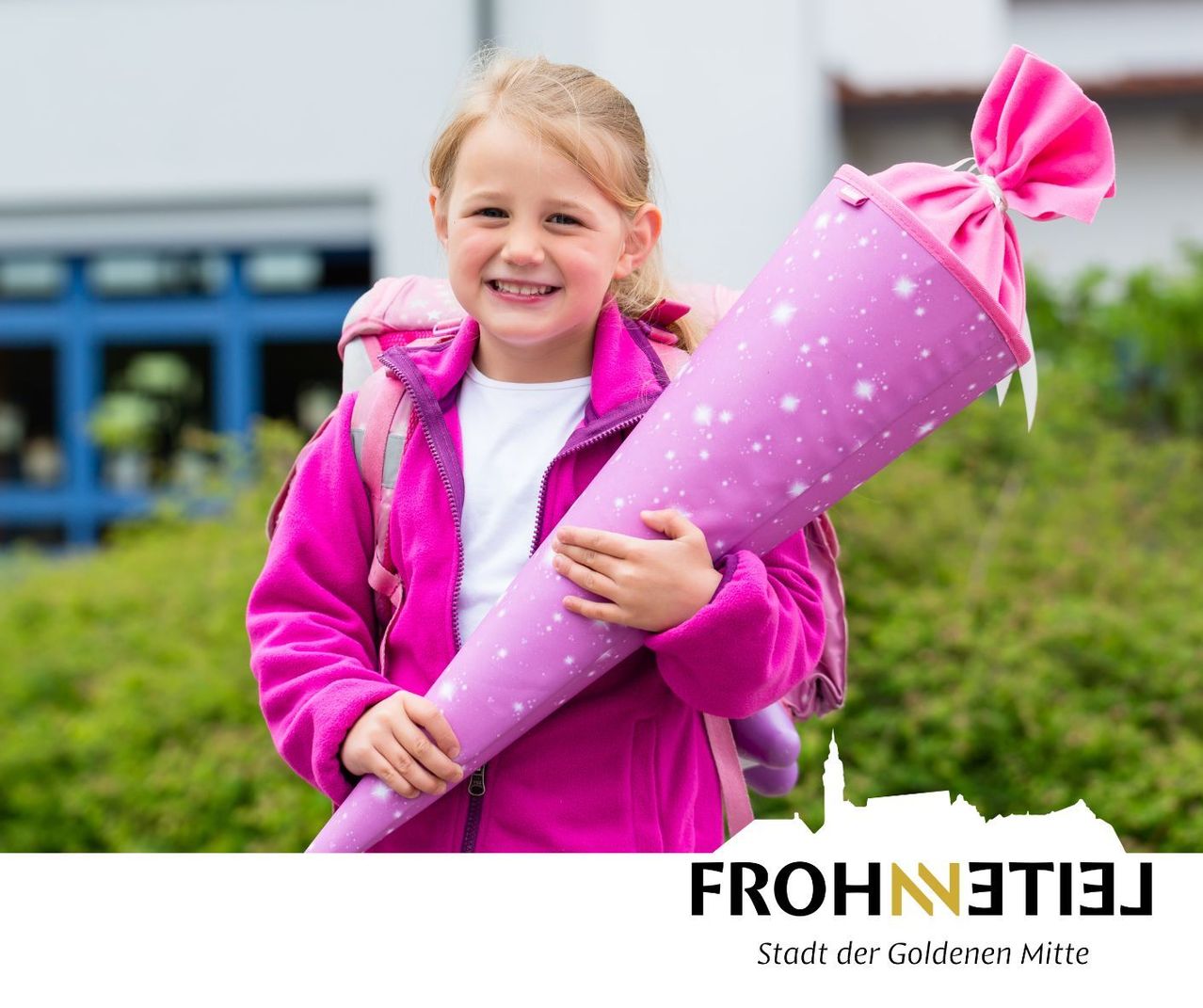 A young girl wearing a pink jacket and carrying a pink backpack is holding a pink pencil case with a bow. She is smiling and standing in front of a building.