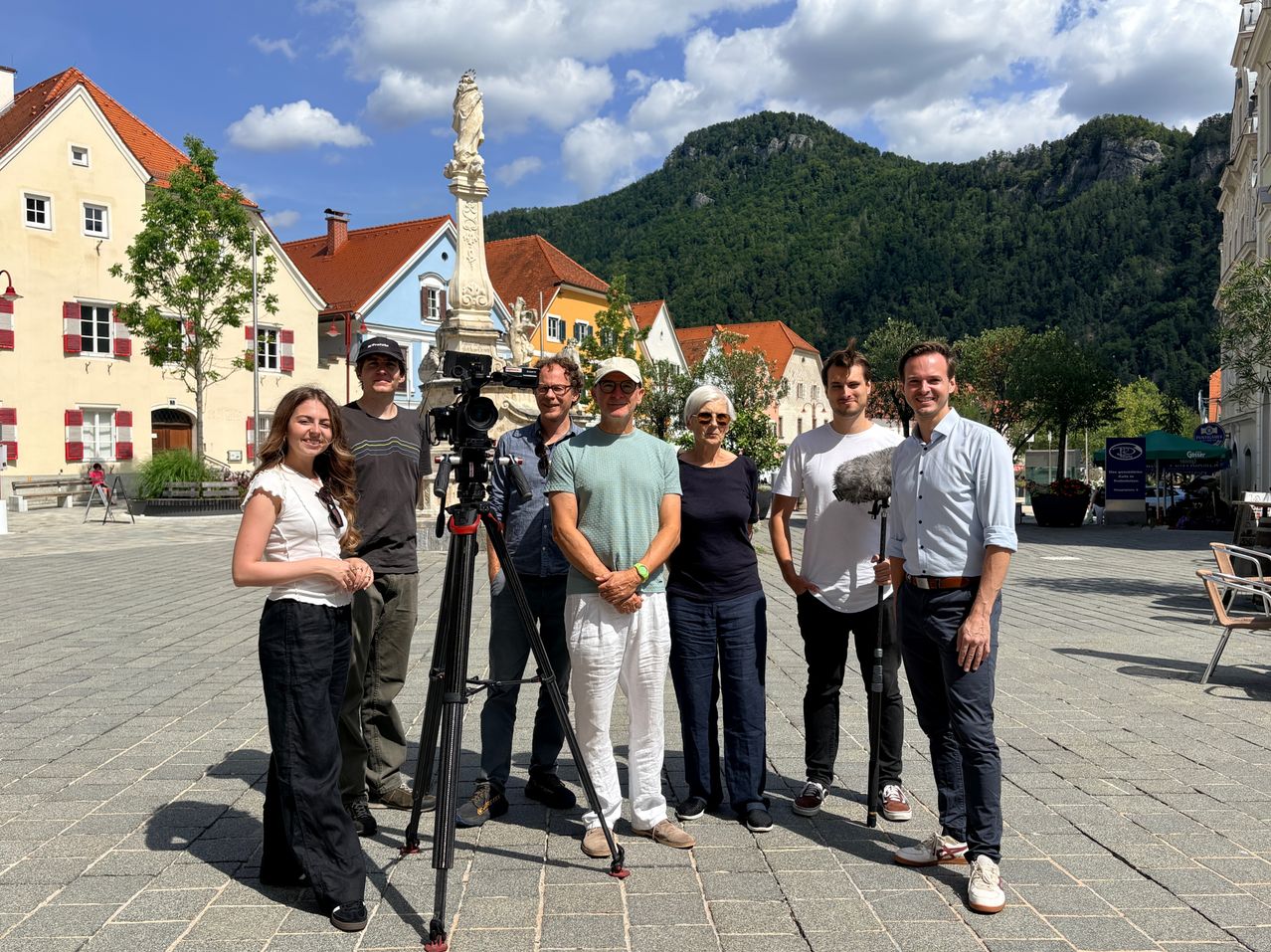 Eine Gruppe von Menschen steht vor einer Kamera auf einem Stativ und lächelt für ein Foto. Dahinter befindet sich ein Denkmal mit einer Statue, und im Hintergrund sind Häuser mit roten Dächern und ein Berg zu sehen.